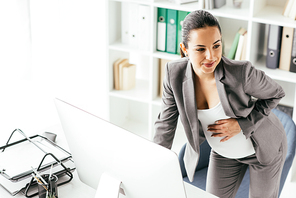 pregnant woman in suit holding belly while standing near table, working on computer and looking away