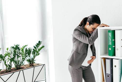 pregnant woman enduring pain and leaning on bookcase in office