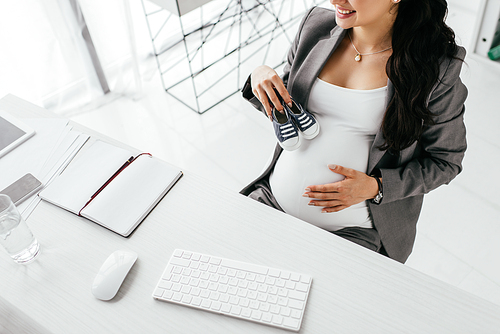 cropped view of pregnant woman sitting behind table with computer keyboard and mouse and holding small gumshoes