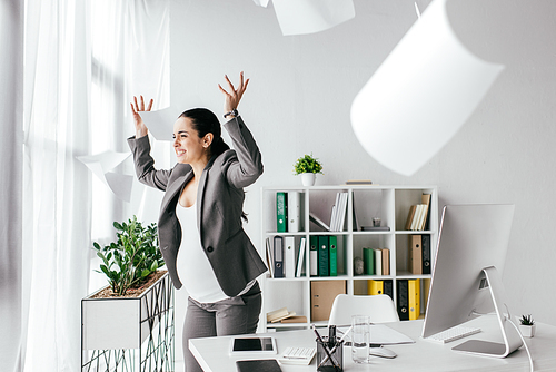 irritated pregnant woman throwing papers in air while standing near table in office