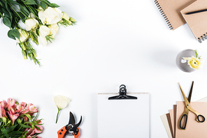 top view of clipboard, secateurs, flowers, notebooks, scissors and craft paper on white