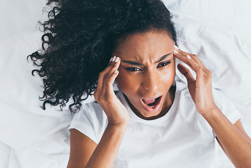 exhausted african american woman screaming while suffering from headache in bed