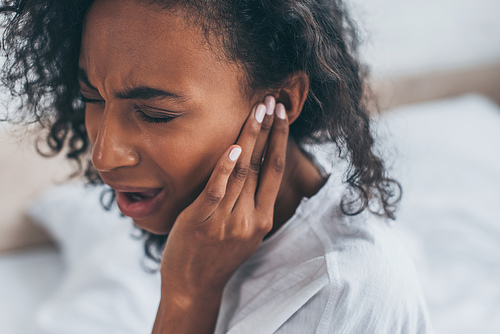 young african american woman suffering from ear pain in bedroom with closed eyes