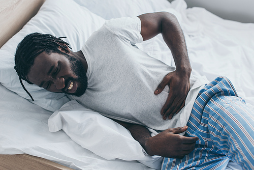 unhappy african american man suffering from abdominal pain while lying in bed