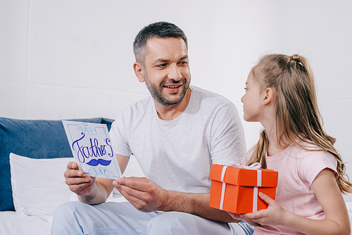 adorable daughter sitting with gift box near happy dad holding fathers day greeting card