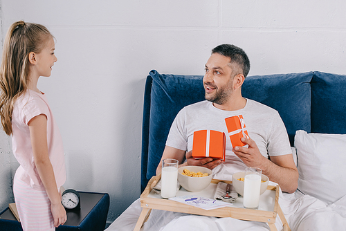 happy man holding fathers day gift boxes while having breakfast in bed near adorable daughter