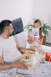smiling man holding tray with breakfast while sitting in bed near adorable daughter covering face with fathers day greeting card