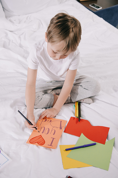 cute boy making fathers day greeting card with i love you dad inscription and heart symbol while sitting on bedding