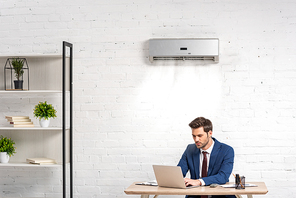 handsome businessman using laptop while sitting at workplace under air conditioner