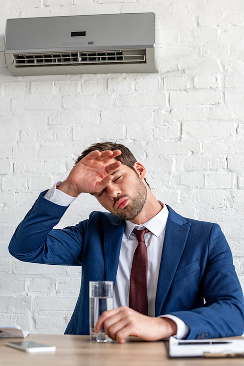 young businessman suffering from heat while sitting under air conditioner with glass of water