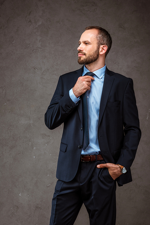 handsome businessman standing with hand in pocket and touching tie on grey