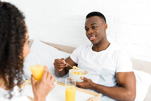 selective focus of african american man with breakfast bowl looking at girlfriend, lying in bed