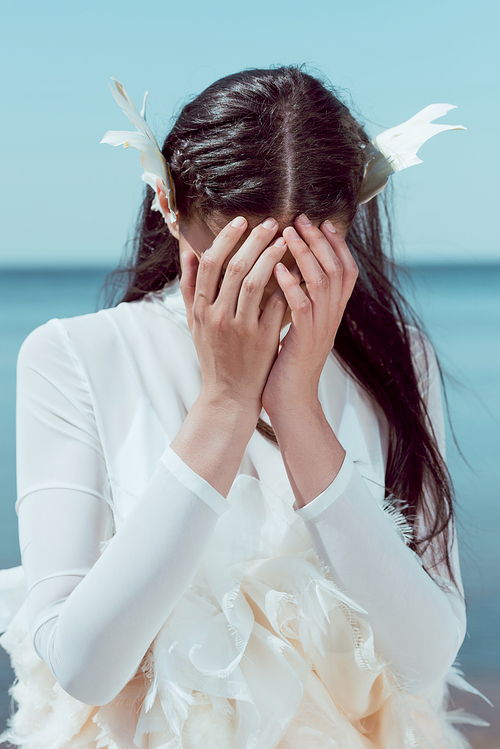 upset woman in white swan costume standing on blue river and sky background, covering face by hands