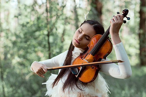 adult woman in white swan costume standing on forest background, playing on violin