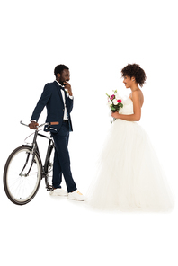 african american bridegroom standing near bicycle and looking at cheerful bride with flowers isolated on white