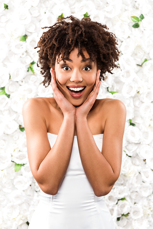 excited african american bride touching face while  near flowers