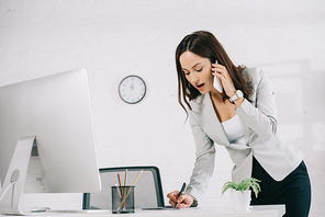 attentive secretary talking on smartphone and writing in notebook while standing at workplace