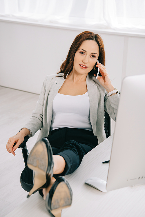 thoughtful, elegant secretary talking on smartphone while sitting with legs on desk