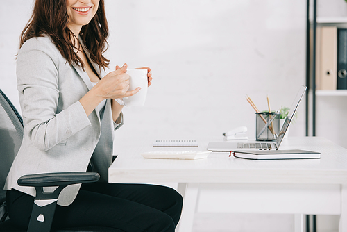 cropped view of smiling secretary holding coffee cup while sitting at workplace in office