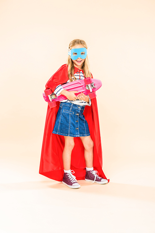 full length view of child in mask and hero cloak holding skateboard on pink