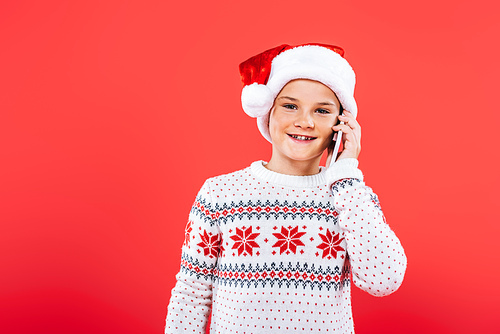 front view of smiling kid in santa hat talking on smartphone isolated on red