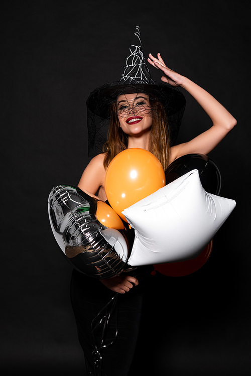 happy woman touching witch hat near balloons on black