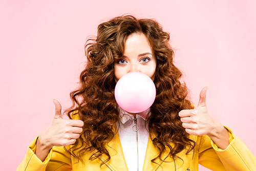 attractive happy curly girl with bubble gum showing thumbs up, isolated on pink