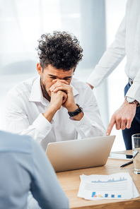 Selective focus of businessman pointing at laptop to sad african american colleague in office