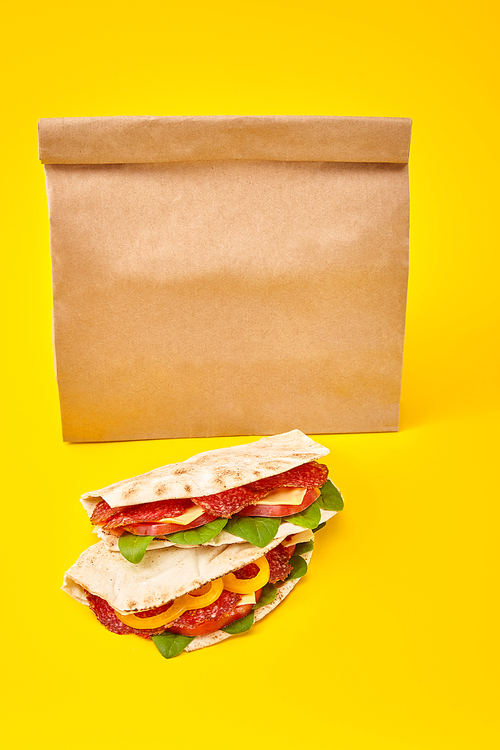 fresh sandwiches with salami, pita, vegetables and cheese on yellow background near paper takeaway bag