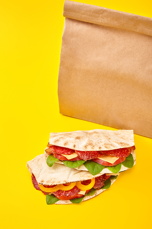 fresh sandwiches with salami, pita, vegetables and cheese on yellow background near paper takeaway bag