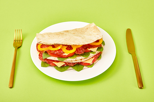 fresh sandwich with salami, pita, vegetables and cheese served on plate near golden fork and knife on green background