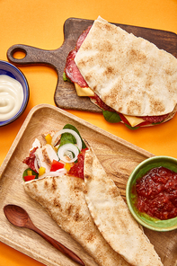 top view of fresh sandwich with salami in pita and burrito with chicken on boards near sauces on orange background