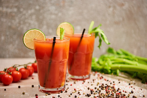 bloody mary cocktail in glasses with straws and lime near salt, pepper, tomatoes and celery on grey background