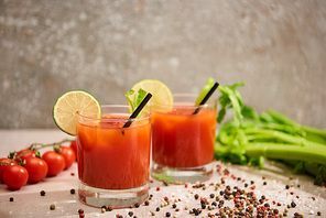 bloody mary cocktail in glasses with straws and lime near salt, pepper, tomatoes and celery on grey background