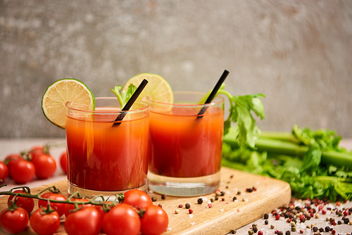 bloody mary cocktail in glasses with straws and lime on wooden board near salt, pepper, tomatoes and celery