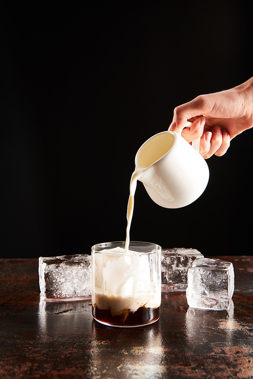 cropped view of woman poring milk in glass with ice cubes isolated on black