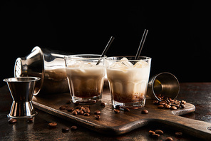 white russian cocktail in glasses with straws on wooden board with coffee grains and shaker isolated on black