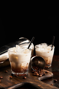 white russian cocktail in glasses with straws on wooden board with coffee grains and shaker isolated on black