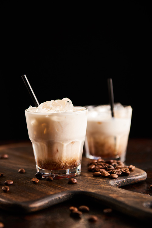 white russian cocktail in glasses with straws on wooden board with coffee grains isolated on black