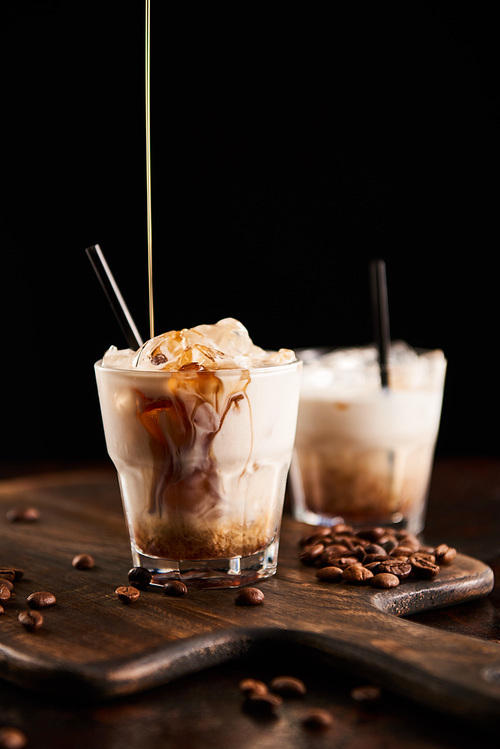 coffee liquor pouring into white russian cocktail in glasses with straws on wooden board with coffee grains isolated on black