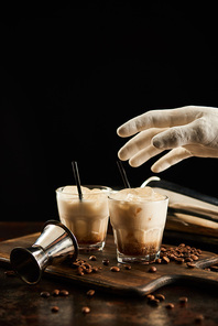 decorative hand above white russian cocktail in glasses with straws on wooden board with coffee grains isolated on black
