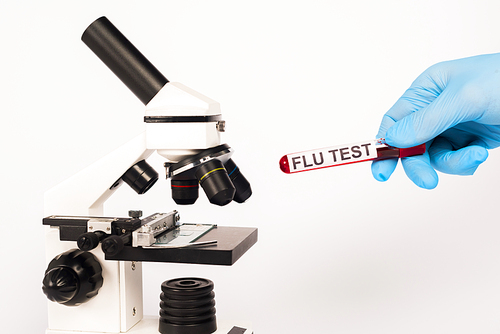 cropped view of scientist in latex glove holding test tube with flu test lettering near microscope isolated on white