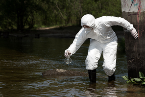 water inspector in protective costume, respirator and goggles holding flask with water sample