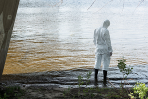 back view of water inspector in protective costume holding flask while standing in river