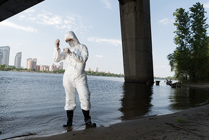 full length view of water inspector in protective costume and respirator taking water sample at river