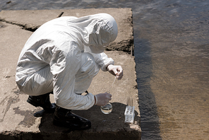 water inspector in protective costume, latex gloves and respirator taking water sample at river