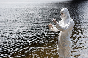 water inspector in protective costume, latex gloves and respirator taking water sample at river