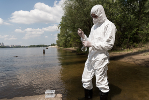 water inspector in protective costume, latex gloves and respirator taking water sample at river