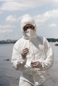 front view of water inspector in protective costume, latex gloves and respirator holding flask with water sample at river