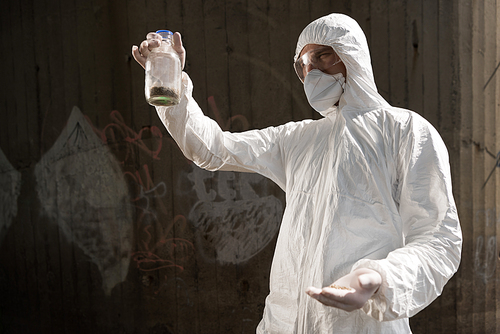 ecologist in protective costume and respirator holding bottle with soil sample